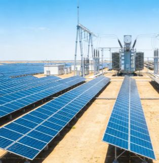 A real-world view of a large-scale, centralized ground-mounted solar photovoltaic power station. On a vast sandy plain, long arrays of deep blue solar panels stretch into the distance. At the far right of the array's end, a giant industrial high-voltage transformer is installed, connected to a dense network of power transmission cables and supported by a towering metal gantry structure. To the left is a white grid-connected housing, set against a clear blue sky, comprehensively showcasing the industrial scene of grid-level photovoltaic power generation and voltage boosting systems.