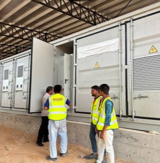 Inside an industrial plant with a large steel-structured roof and earthen platform, a scene of on-site installation and operation of a containerized battery energy storage system is displayed. In the center of the image are neatly arranged large, standard white energy storage containers, securely placed on concrete bases. Yellow lightning bolt high-voltage danger triangle warning signs are printed on multiple inspection doors of the containers, and the right-side panel integrates large metal ventilation louvers. At this moment, one of the container's doors is fully open, revealing the gray-white internal control cabinet. Four male workers are gathered in front of the equipment: two are standing in front of the open door inspecting or discussing the equipment, while the other two young technicians, wearing bright yellow fluorescent safety vests, are talking on the right. The overall scene showcases the tense but orderly construction and commissioning atmosphere at an industrial-grade new energy storage project site.