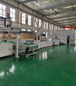 This is a real-life photograph showcasing the interior of a modern energy storage system manufacturing base. The main focus is a long, parallel, white, automated intelligent assembly line that stretches across the entire workshop from left to right: the left side features an automatic feeding mechanism with a green conveyor belt and a yellow metal safety mesh frame; the middle section is an automated processing area composed of continuous white enclosed cabinets; and at the right end is a tall, independent white central control and monitoring cabin with an embedded intelligent LCD display screen for monitoring production data. The workshop floor is entirely covered with a glossy, mirror-like dark green epoxy floor paint that clearly reflects the equipment. The building structure is grand, with several tall, multi-pane glass windows on one side, and neatly arranged industrial lighting fixtures and longitudinally extending overhead crane beams mounted on the towering steel ceiling. The entire workshop environment is clean, tidy, and orderly, demonstrating high standards of industrial manufacturing.