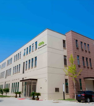 A photograph of the exterior of a modern corporate headquarters building against a clear, high, pure blue sky. The main subject is a multi-story modern industrial park building, framed by a diagonal perspective: the left side of the building features a large, elegant off-white facade with rows of narrow, dark-colored glass windows arranged in a staggered pattern. A transparent glass entrance is located on the ground floor, with several potted green plants neatly placed in front. In a prominent position on the upper right of the off-white facade, a three-dimensional brand logo composed of two connected green mountain peaks or an infinity symbol is embedded, with the black English word 'GreenMore' below. The right side of the building features a stable, reddish-brown rectangular panel, with a young, lush ginkgo tree planted at its base. The rear of a black SUV is parked on the concrete ground in the lower right corner. Under the bright sunlight, the entire building is clearly defined, clean, and presents the fresh image of a modern, independently operated base for a new energy technology company.