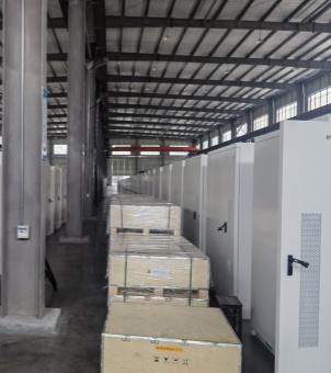 This is a real-life photo taken inside a large, spacious, modern industrial plant, showing the mass production and packaging of high-voltage stacked energy storage units, ready for shipment. The image uses a deep perspective composition: on the right, a row of tall, white steel integrated energy storage cabinets stretches vertically and is neatly arranged. Each cabinet has a strip of metal ventilation mesh on the side and a black rotary lock handle on the front. The cabinets seem to stretch as far as the eye can see, showcasing the scale of mass production. On the left side of the cabinet array, in the central aisle, several heavy-duty wooden crates and pallets are neatly stacked, their surfaces wrapped in transparent plastic moisture-proof wrapping film and reinforced with dense green strapping. To the left of the plant is a row of towering gray I-beam structural columns. The high ceiling features a regular, wave-shaped steel structure design, with rows of industrial lighting chandeliers installed along its length. In the distance, high-level skylights and red overhead crane beams are faintly visible. The entire workshop floor is a clean, gray, hardened cement floor, presenting a panoramic view of the orderly and fully operational industrial scene of the new energy manufacturing base, with large-scale production and delivery at full capacity.