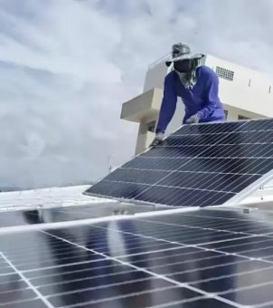 This is a real-life photo showcasing the on-site installation of a solar photovoltaic system on a building's flat roof. The image is shot from a low angle, with the foreground featuring a large, horizontally laid-out matrix of dark black monocrystalline silicon solar panels, their surfaces slightly reflective in the weak sunlight, their silver-white conductive grid lines clearly visible. In the mid-ground, a construction worker wearing a blue long-sleeved work uniform, a camouflage hat, a full-face mask, and professional non-slip gloves stands bent over behind a tilted metal mounting bracket, steadily supporting a large rectangular solar photovoltaic panel that is about to be secured. The background is a white, modern residential or commercial building high above, with a white, overcast sky covered by thick clouds. The entire scene is realistic and natural, vividly demonstrating the engineering details of the on-site construction and core component assembly of the distributed rooftop photovoltaic power generation system.