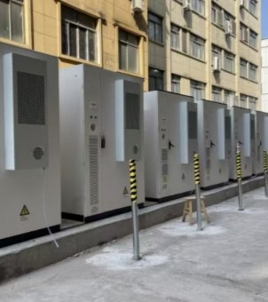 This is a real-world photograph showcasing a commercial and industrial energy storage system's outdoor distributed rack array deployment project. The image uses a perspective composition from near to far: a row of six to seven tall, white, rectangular energy storage racks are lined up, securely mounted on a raised, flat gray concrete pedestal in a factory or industrial park. Each rack has a tall, independent gray air conditioning cooling unit mounted externally or internally on its front side or front door. The unit's surface features a high-density rectangular metal heat dissipation mesh and a circular fan exhaust vent below. A triangular yellow high-voltage hazard warning sign is affixed to the lower left corner of the foremost rack. Extending along the concrete ground in front of the energy storage rack array, silver metal crash barriers are vertically installed at intervals. The upper middle section of these barriers is covered with prominent yellow and black diagonally striped safety reflective tape. A simple wooden stool is placed at the edge of the pedestal in the middle of the rack group. The background is a pale yellow, multi-story industrial or modern office building with densely packed, multi-pane windows. Under bright natural light, the entire scene showcases the application of an efficient and standardized industrial and commercial multi-energy complementary energy storage infrastructure project.
