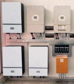 This is a real-life photograph showcasing a standardized wall-mounted installation of a home energy storage battery and hybrid inverter system. The image depicts a meticulous electrical layout against a white wall: on the left are three clean, white rectangular wall-mounted energy storage battery modules (one in the upper left and two side-by-side in the lower left), each with a gray decorative line at the bottom front and a small LCD digital status display. In the upper right are two gray rectangular hybrid inverters, each with a circular control panel and display window at the center. The rightmost inverter has a safety warning label with yellow and black diagonal stripes on its lower casing. Thick red and black DC cables extend from the bottom of all devices, neatly and vertically connecting to a silver metal cable tray running horizontally through the center of the image. In the lower right, a row of black rectangular circuit breaker fuse units is embedded under the metal cable tray, below which hangs a striking bright orange metal distribution control box, with four white vertical conduits extending from its bottom. The entire system was installed neatly and fit together perfectly, fully demonstrating the professional assembly process of a modern household clean energy backup power system.