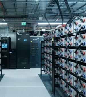 This is a real-life photograph showcasing the backup energy storage and server rack layout inside a modern telecommunications or Internet Data Center (IDC). The image employs a deep perspective composition extending from the right: in the foreground, a row of five-story-high open black metal battery racks displays dozens of densely packed, rectangular, gray-white dedicated communication backup batteries. Each battery has prominent red and blue circular terminals on its front, connected in series and parallel by robust black insulated cables. In the mid-ground and background, on a gray anti-static floor, stands a row of standard black IT server racks and tall UPS power control cabinets. Some racks have transparent glass or mesh windows on their front doors, subtly displaying cyan-blue digital status indicator lights. The ceiling of the server room is completely open, densely packed with intricate silver-gray mesh cable trays and thick main power cable bundles. Above, green emergency exit signs with white running figures hang. The entire space is brightly lit, exuding a high-density sense of electrical technology and a rigorous industrial order.