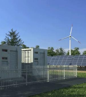 This photograph, taken on a clear day, showcases the industrial application of a modern integrated wind, solar, and energy storage microgrid and off-grid power supply system. The image employs a multi-layered perspective composition: in the left foreground, a heavy-duty energy storage system, protected by a white metal safety fence, is erected at the edge of a green lawn. It consists of two gray-white grid-grade energy storage container cabinets, with robust horizontal reinforcing ribs and industrial ventilation louvers on their exteriors. In the midground, behind a buffer zone, a large, neatly arranged array of blue solar photovoltaic panels faces the sun at an angle. In the right background, behind a dense green forest, stands a towering, modern, three-bladed white wind turbine, its three rotating blades tipped with red warning stripes. Above the entire scene is a clear, cloudless blue sky, and the ground consists of clean asphalt roads, neatly trimmed lawns, and trees, clearly outlining a picture of a distributed microgrid's multi-energy complementary green and clean energy infrastructure.