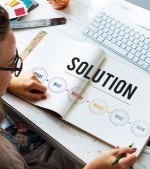 This is a top-down photograph showcasing a designer brainstorming system design solutions at their desk. On the left is the profile of a woman with black-rimmed glasses and her hair tied back, leaning over a wooden desk. Her left hand rests on a fully open, light yellow grid notebook, while her right hand holds a green pencil. The notebook's right page displays the large, bold, black word 'SOLUTION' underlined. On the left, from the top left to the bottom right, are six light-colored, gradient circular labels connected by dotted lines. Each label contains the 5W1H analysis method: blue 'WHAT', purple 'WHY', red 'WHERE', yellow 'WHEN', blue-gray 'WHO', and green-gray 'HOW'. Above the notebook, a white, flat wireless keyboard is neatly arranged on the desk. Scattered to the upper left of the notebook are a black stainless steel ruler, a small black rectangular tool, and a colorful design swatch with its edge peeking out. The entire scene is lit with soft, natural light and a warm color scheme, creating a professional atmosphere conducive to rigorous thinking in office and customized project design.