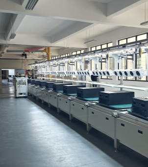 This is a photograph taken from a deep, oblique perspective, showcasing a modern, automated assembly and testing line for new energy commercial and industrial inverters or energy storage equipment in an industrial workshop. The image employs a strong sense of depth and oblique perspective: an extremely long transmission line, constructed of stainless steel and green anti-static conveyor belts, extends infinitely from the right front to the left rear. More than ten black rectangular industrial converter cabinets (PCS or hybrid inverters) are neatly and evenly arranged side-by-side on the line, undergoing assembly or testing. The cabinet panels reveal precise wiring holes and fixing screws. Above the line is a long, silver metal suspension bracket, on which hangs a dense, neat row of pure white, long, strip-shaped digital testing instruments or monitoring components. Each component has four black, round sensor or lens markings side-by-side on its front, with black power cables densely arranged above them. A long, high-brightness fluorescent light strip at the top of the bracket provides bright illumination for each workstation. At the far left end of the assembly line stands a mobile, digital central control testing platform equipped with a monitor, keyboard, and multi-layered cabinets. In the background, other workshop aisles and some desks and chairs are faintly visible. The entire factory is spacious, featuring a white concrete load-bearing beam and column structure and a long row of bright, black-framed glass windows. The overall environment is clean and professional, fully demonstrating the large-scale, high-standard intelligent industrial manufacturing and stringent quality control of new energy electrical equipment.