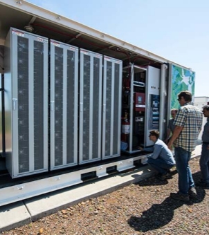 An outdoor photograph taken on a clear day shows engineers from a communications and microgrid developer conducting technical testing on a containerized energy storage system at a project site. The image employs a perspective composition extending from left to right: on the left is a tall, spacious white standard heavy-duty container, its long side doors fully opened or removed, revealing extremely compact and sophisticated industrial electrical equipment inside. Inside the container, five tall, silver-gray, multi-layered modular energy storage battery cabinets stand neatly and tightly side-by-side from left to right, their doors featuring fine, continuous ventilation grilles and long silver metal handles. To the right of the battery cabinets, a complex power distribution and control system is tightly nested, with multiple thick red power cables visible, and a red cylindrical fire extinguishing gas cylinder is also present. In front of the open electrical compartment on the right side of the container, a young technician wearing a blue long-sleeved shirt and blue jeans is facing the cabinets, crouching on the ground, debugging the wiring. Three other men stood to his right and behind him. The one in the foreground was tall, wearing a blue-green plaid short-sleeved shirt and dark jeans, and sunglasses, watching the commissioning process intently. The two men behind him were also dressed casually, focused on their work. The power station ground was an outdoor wasteland covered with fine gravel and soil, and the container shell on the right was decorated with green and white geometric natural-style mural graffiti. The entire scene was brightly lit, with strong contrasts of light and shadow, clearly presenting the delivery and professional technical support site of the new energy microgrid project.