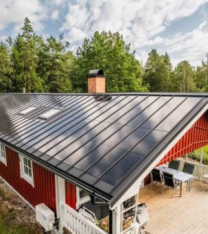 A stunning photograph taken on a clear day, from a bird's-eye view, showcasing a modern Scandinavian-style redwood house featuring a BIPV (Building Integrated Photovoltaic) roof system. The visual focus is a large, sloping black metal standing seam photovoltaic roof. Multiple vertical metal seam lines extend neatly and parallel from the ridge to the eaves, evenly dividing the roof surface into long, continuous strips. The roof surface is remarkably smooth and has a mirror-like, high-end texture, clearly reflecting the azure sky and large white cumulus clouds above. Two rectangular skylights with white frames are located on the upper left side of the roof. A traditional rectangular chimney made of red brick, topped with a black windproof cap, stands at the center of the ridge. Below the roof is a Scandinavian-style house, its exterior walls constructed of striking rust-red vertical wooden slats, complemented by elegant white-framed lattice windows. Extending to the lower right is a spacious outdoor wooden terrace, furnished with a rectangular dining table and several modern black armchairs, surrounded by a white wooden railing. Behind and around the house grows a dense, tall, lush forest of coniferous and broad-leaved trees, above which lies a sunny sky dotted with fluffy white clouds. The entire scene perfectly blends natural ecology, traditional architectural aesthetics, and cutting-edge distributed photovoltaic technology, creating a warm and inviting atmosphere befitting a modern, green, and low-carbon residence.
