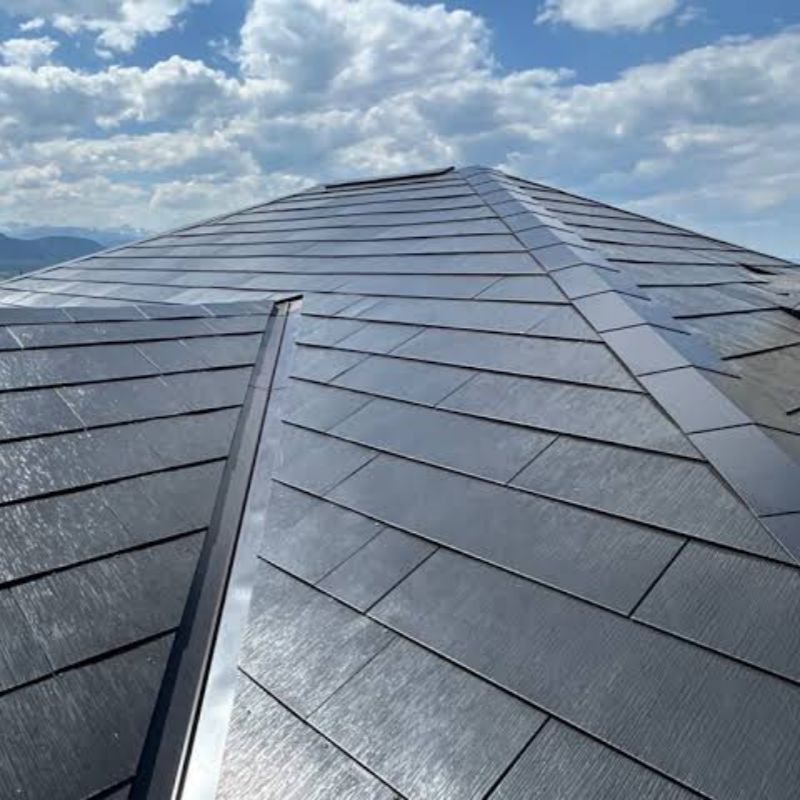 This is a detailed close-up photograph taken from a slightly upward angle, showcasing a multi-sloped roof in a modern residential building equipped with high-end flat solar tiles. The main visual element is the multi-sloped roof, formed by the convergence
