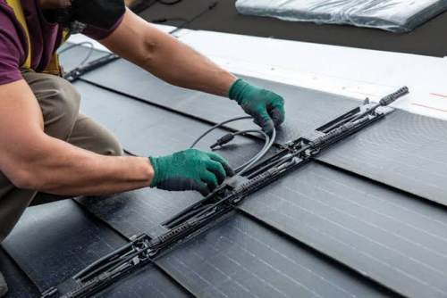 A close-up, realistic photograph shows an installer wearing green non-slip work gloves and brown work pants kneeling on a black roof, carefully arranging and connecting gray cables and waterproof connectors between the photovoltaic roof tiles. The black solar photovoltaic tiles are neatly arranged on the roof, their slotted structure and internal wiring channels clearly visible.